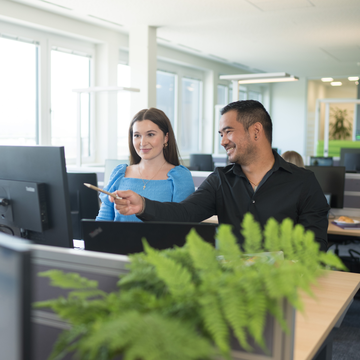 Ein Lehrling und ein Mitarbeiter sitzen an einem Schreibtisch in einem modernen Büro und besprechen Informationen auf einem Computerbildschirm. Im Hintergrund sind weitere Arbeitsplätze und Pflanzen sichtbar, die eine angenehme Arbeitsatmosphäre schaffen.