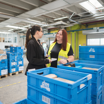 Zwei Mitarbeiterinnen im Verteilzentrum stehen zwischen blauen Lagerkisten und besprechen etwas miteinander. Im Hintergrund sind weitere Kisten auf Paletten zu sehen. Die Mitarbeiterinnen wirken motiviert und fröhlich.