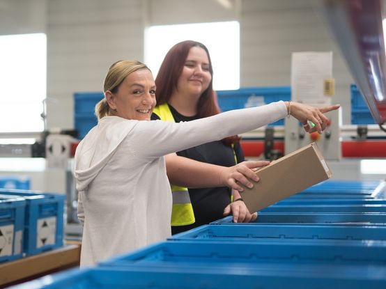 Zwei Mitarbeiterinnen stehen neben dm Kisten und räumen Produkte ein. Eine Kollegin zeigt der anderen Kollegin etwas. Die Stimmung wirkt fröhlich.