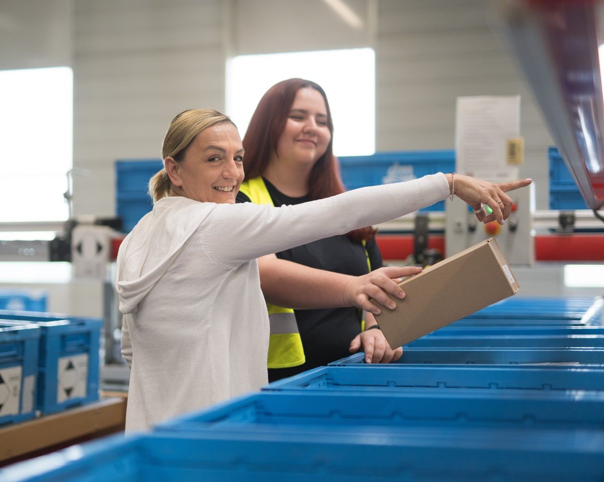 Zwei Mitarbeiterinnen stehen neben dm Kisten und räumen Produkte ein. Eine Kollegin zeigt der anderen Kollegin etwas. Die Stimmung wirkt fröhlich.