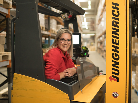 Eine Mitarbeiterin sitzt auf einem Gabelstapler zwischen sehr hohen Lageregalen und lächelt in die Kamera.