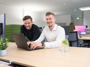 Zwei Mitarbeiter in einem modernen Büro, die lächelnd auf einen Laptop schauen, während ein Glas Wasser mit Minze und Zitrone auf dem Schreibtisch steht.