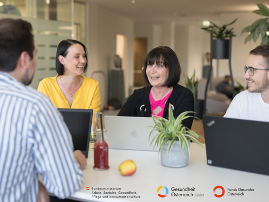 Vier Kollegen sitzen lächelnd um einen Tisch mit Laptops, einer Pflanze und Getränken während einer Besprechung im Büro.