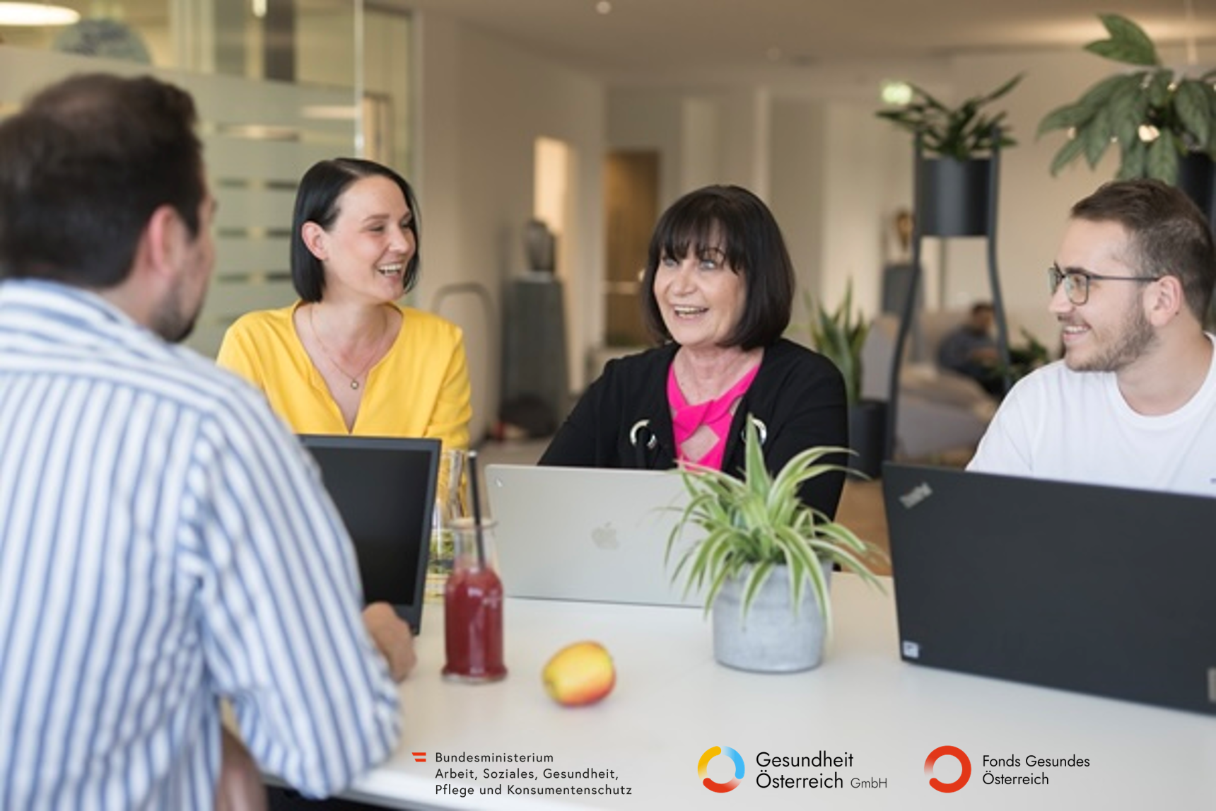 Vier Kollegen sitzen lächelnd um einen Tisch mit Laptops, einer Pflanze und Getränken während einer Besprechung im Büro.