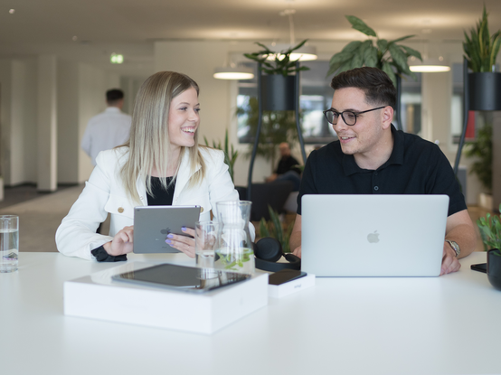 Eine Mitarbeiterin und ein Mitarbeiter sitzen nebeneinander an der Highbench. Er hat einen Laptop vor sich, während sie ein Tablet nutzt. Im Hintergrund ist der Cafébereich zu sehen.