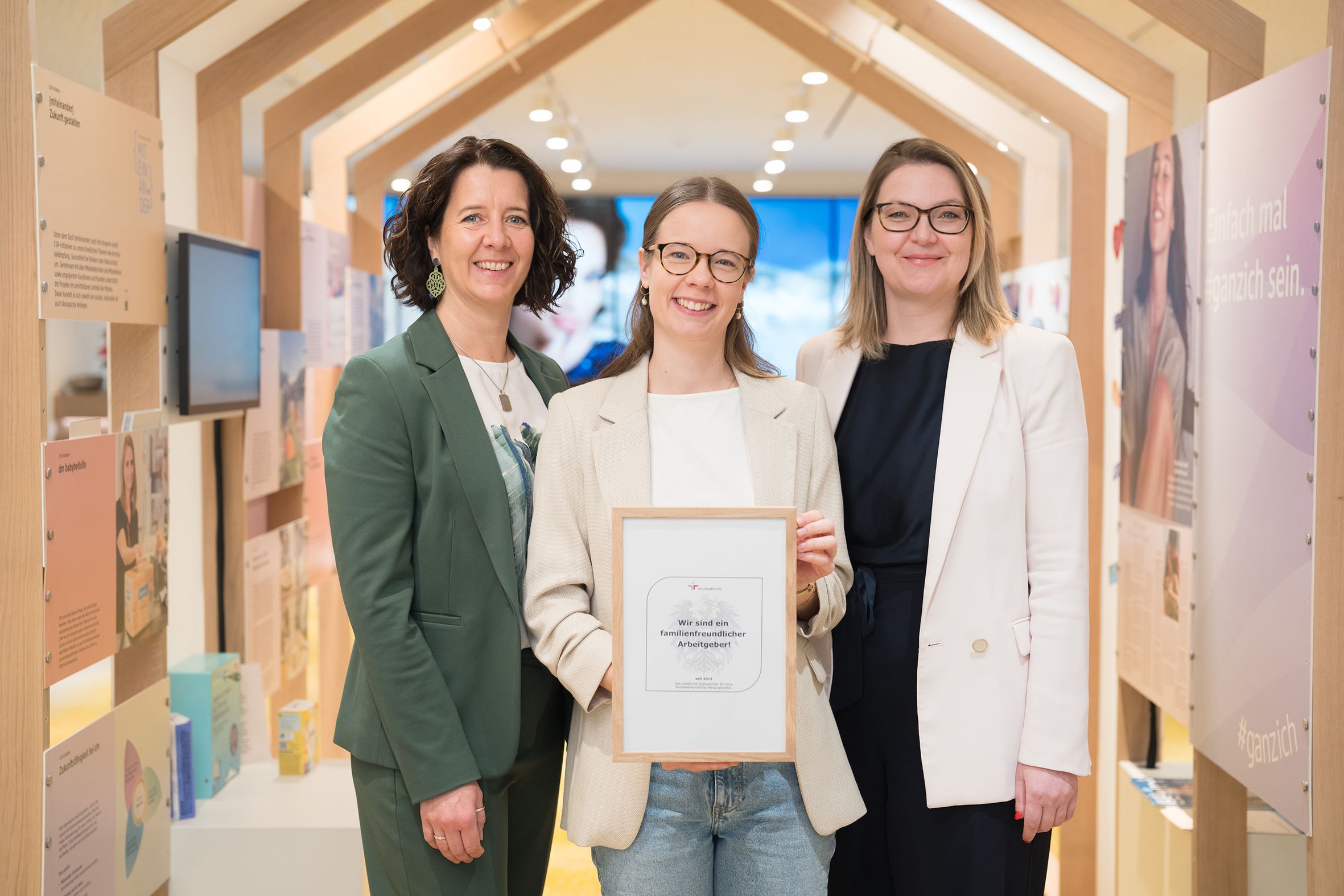 Drei Frauen stehen nebeneinander im Empfangsbereich des dm dialogicum. Die Frau in der Mitte hält ein Zertifikat und alle lächeln..