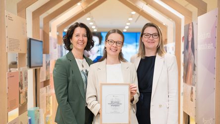Drei Mitarbeiterinnen lächeln in einem modernen, gut beleuchteten Raum. Die Frau in der Mitte hält ein Zertifikat in einem hölzernen Rahmen.