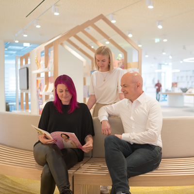 Drei Personen sind gemeinsam im Empfangsbereich des dm dialogicum. Eine sitzende Frau liest ein Magazin, während der sitzende Mann daneben mit in das Magazin schaut. Eine weitere Frau steht hinter den beiden und schaut ebenfalls in das Magazin.