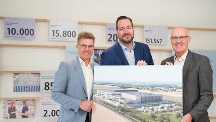 Drei Männer in Anzügen halten eine große Architekturzeichnung eines modernen Gebäudes in den Händen und lächeln.