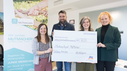 Vier Personen stehen vor einem Banner auf dem Mutmachen steht. Sie halten einen Bild mit einem Spendenbetrag in der Hand.
