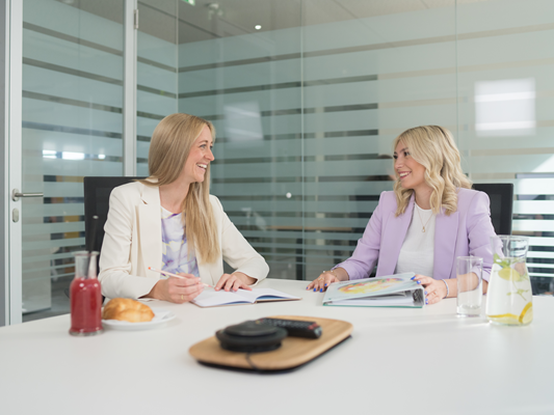 Zwei Mitarbeiterinnen sitzen an einem Tisch in einem modernen Besprechungsraum und unterhalten sich über Unterlagen. Auf dem Tisch befindet sich Verpflegung und Arbeitsmaterialen. Sie zeigen Begeisterung und Freude an ihrer Tätigkeit.