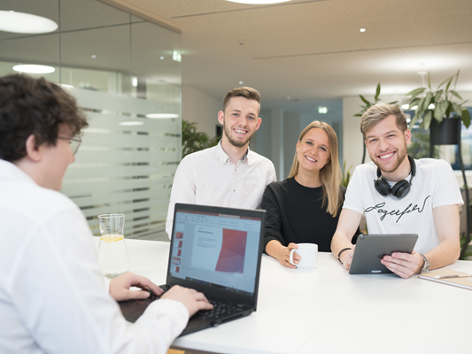 Vier Personen in einem modernen Büro, drei lächeln in die Kamera, eine arbeitet an einem Laptop. Auf dem Tisch stehen ein Tablet und ein Glas Wasser.