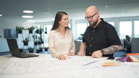 Ein Mitarbeiter und eine Mitarbeiterin stehen an einer Highbench im Büro und arbeiten konzentriert an Filialplänen, die vor ihnen ausgebreitet sind.