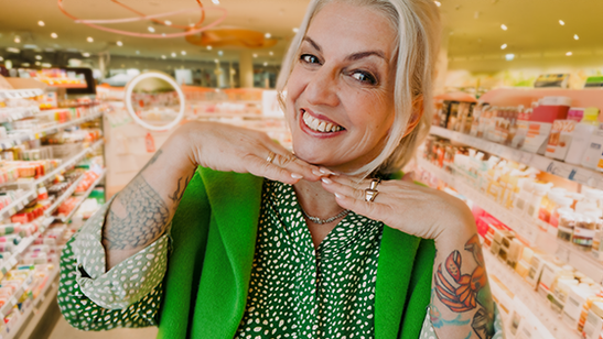 Eine Mitarbeiterin posiert in einem modernen dm Markt zwischen Regalen und lächelt in die Kamera. Auf ihren Armen sind Tattoos zu sehen und die Stimmung wirkt sehr ansprechend und locker.