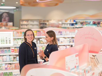 Zwei Mitarbeiterinnen in einem dm Markt stehen in der Kosmetikabteilung und unterhalten sich über ein Produkt. Beide wirken freundlich.