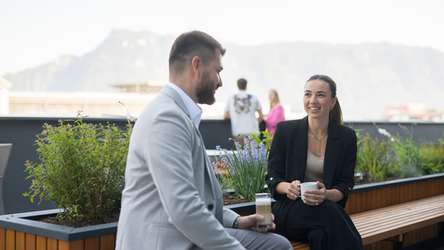 Eine Mitarbeiterin und ein Mitarbeiter sitzen mit einem Kaffee auf der Terrasse in der dm Zentrale. Die beiden unterhalten sich und haben Spaß. Im Hintergrund sind weitere Mitarbeitende zu sehen und einige Pflanzen.