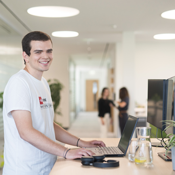 Ein Mitarbeiter steht an einem Hochtisch und arbeitet an einem Laptop. Im Hintergrund sind Kolleginnen zu sehen und ein modernes und helles Büro.