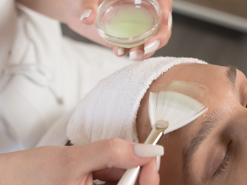 Eine Mitarbeiterin trägt eine Gesichtsmaske auf. Sie hält eine kleine Schale mit der Maske in der einen Hand und verwendet einen Pinsel, um die Maske gleichmäßig aufzutragen.