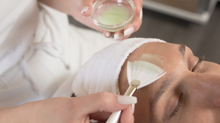 Eine Mitarbeiterin trägt eine Gesichtsmaske auf. Sie hält eine kleine Schale mit der Maske in der einen Hand und verwendet einen Pinsel, um die Maske gleichmäßig aufzutragen.