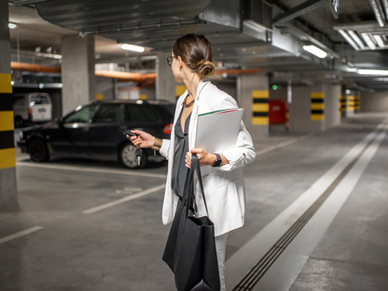 Eine Frau in einem weißen Blazer hält einen Ordner und eine Tragetasche, während sie in einer Tiefgarage geht und zu einem geparkten schwarzen Auto schaut.