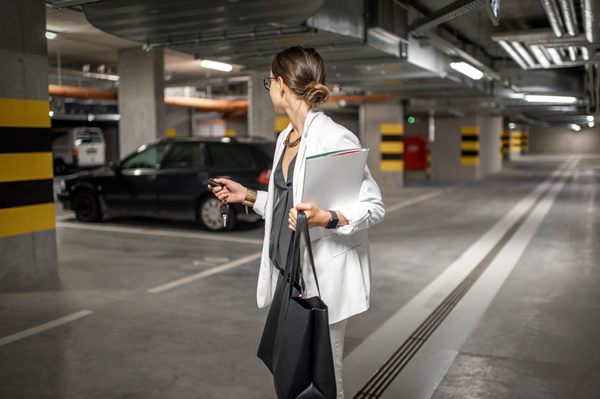 Eine Frau in einem weißen Blazer hält einen Ordner und eine Tragetasche, während sie in einer Tiefgarage geht und zu einem geparkten schwarzen Auto schaut.