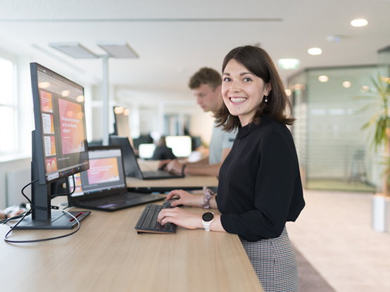 Mitarbeiterin steht auf der Open Space Bürofläche am Schreibtisch und arbeitet, während sie in der Kamera lächelt. Im Hintergrund sieht man einen Kollegen und Besprechungsräume.