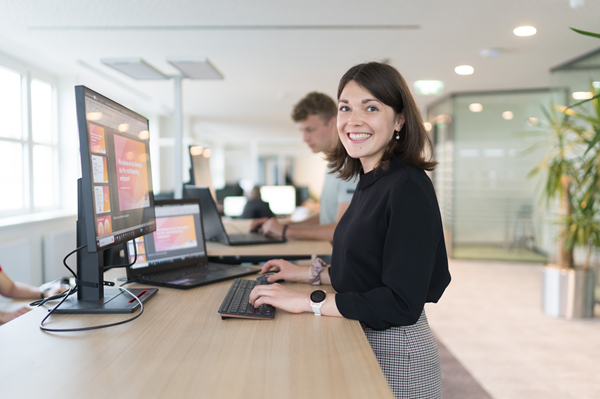 Mitarbeiterin steht auf der Open Space Bürofläche am Schreibtisch und arbeitet, während sie in der Kamera lächelt. Im Hintergrund sieht man einen Kollegen und Besprechungsräume.