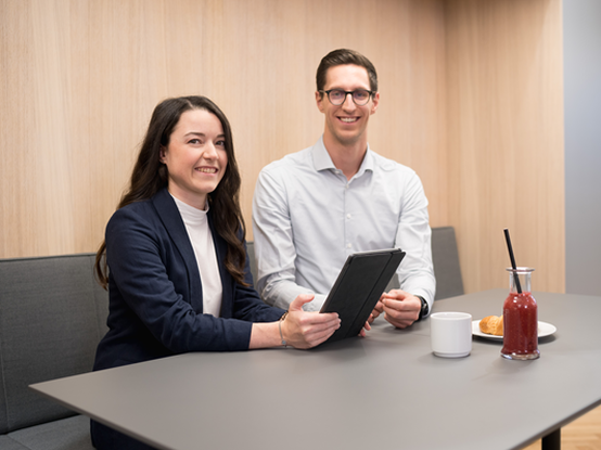 Eine Mitarbeiterin und ein Mitarbeiter sitzen an einem Tisch, lächeln und nutzen ein Tablet. Auf dem Tisch stehen eine Tasse, ein Croissant und ein Smoothie. Im Hintergrund ist eine Wand aus hellem Holz zu sehen, die zur gemütlichen Atmosphäre beiträgt.