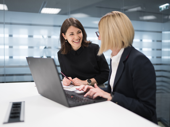 Zwei Mitarbeiterinnen sitzen mit einem Laptop im Besprechungsraum und lachen.