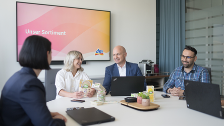 Zwei Mitarbeiterinnen und zwei Mitarbeiter sitzen in einem Besprechungsraum mit Laptops. Sie lächeln und diskutieren miteinander. Auf einem Präsentationsbildschirm steht "Unser Sortiment".