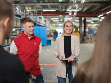 Ein Lehrling und eine Mitarbeiterin stehen im Verteilzentrum und haben mit ihrem Team eine Besprechung.