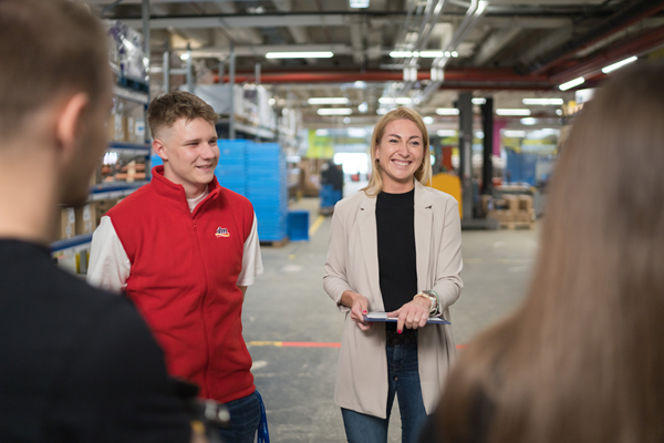 Ein Lehrling und eine Mitarbeiterin stehen im Verteilzentrum und haben mit ihrem Team eine Besprechung.