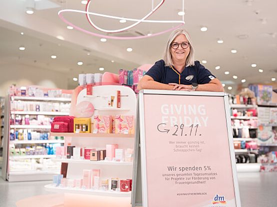 Mitarbeiterin steht im dm Markt und lehnt sich auf einen Aufsteller. Auf dem Aufsteller steht "Giving Friday 29.11.".