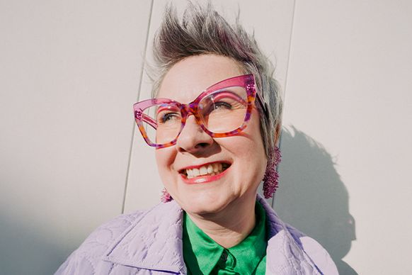 Eine lächelnde Person mit kurzen Haaren, einer großen rosa Brille und einem bunten Outfit steht vor einer hellen Wand im strahlenden Sonnenlicht.