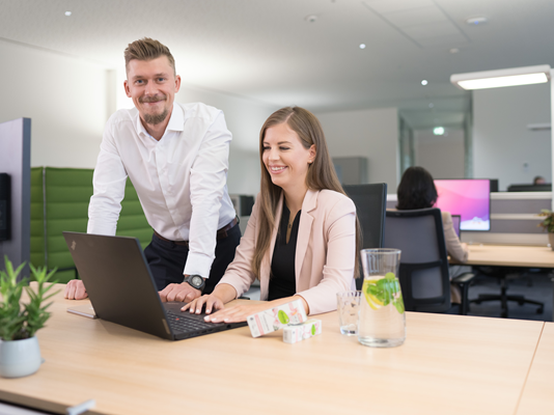 Eine Mitarbeiterin, die am Laptop an einem Schreibtisch sitzt, während ein Mitarbeiter neben ihr steht und in die Kamera schaut. Auf dem Schreibtisch befinden sich dm-Produkte und eine Glaskaraffe, und im Hintergrund sitzt eine Kollegin an einem anderen Schreibtisch.