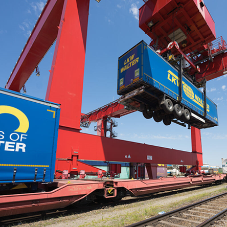 Ein großer roter Kran hebt einen blauen Frachtcontainer auf eine Flachwagen des Zuges in einem Bahnhofsbereich unter einem klaren blauen Himmel.