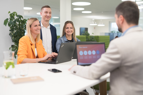 Vier Mitarbeitende in einem modernen Büro um eine Highbench, wobei zwei Mitarbeiterinnen und ein Mitarbeiter mit Laptops sitzen und ein weiterer Mitarbeiter steht, während sie sich angeregt unterhalten. Der Raum ist hell und offen, mit größeren Pflanzen im Hintergrund.