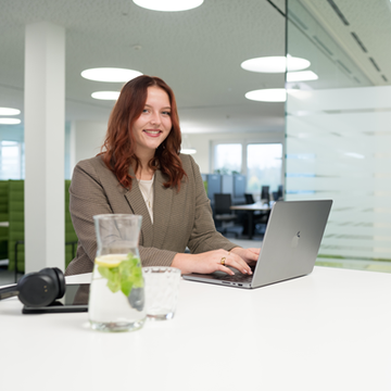 Eine rothaarige Mitarbeiterin sitzt an einem modernen Bürotisch und arbeitet konzentriert an ihrem Laptop. Neben ihr befinden sich ein Tablet, Kopfhörer und ein Glas Wasser.