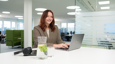 Eine rothaarige Mitarbeiterin sitzt an einem modernen Bürotisch und arbeitet konzentriert an ihrem Laptop. Neben ihr befinden sich ein Tablet, Kopfhörer und ein Glas Wasser.