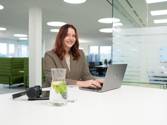 Eine rothaarige Mitarbeiterin sitzt an einem modernen Bürotisch und arbeitet konzentriert an ihrem Laptop. Neben ihr befinden sich ein Tablet, Kopfhörer und ein Glas Wasser.