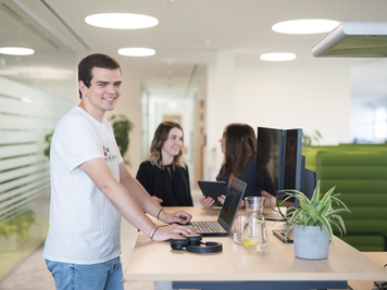 Ein Mitarbeiter arbeitet stehend an einem Schreibtisch am Laptop, während zwei Mitarbeiterinnen im Hintergrund miteinander sprechen.
