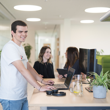 Ein Mitarbeiter arbeitet stehend an einem Schreibtisch am Laptop, während zwei Mitarbeiterinnen im Hintergrund miteinander sprechen.