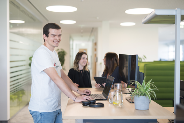 Ein Mitarbeiter arbeitet stehend an einem Schreibtisch am Laptop, während zwei Mitarbeiterinnen im Hintergrund miteinander sprechen.