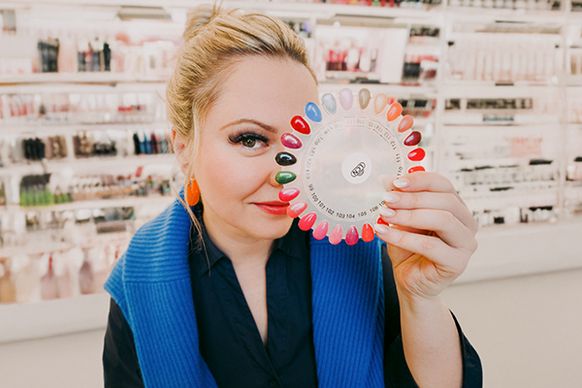 Eine Mitarbeitterin steht in einem Kosmetikstudio vor einem Regal mit verschiedenen Produkten. Die Frau hält einen Farbkreis für Nagellacke hoch. Die Athmophäre wirkt modern und hell.