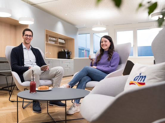 Zwei Mitarbeitende sitzen in einem modernen Café im Büro und unterhalten sich. Ein Mitarbeiter hält ein Tablet, während seine Kollegin eine Tasse Kaffee in der Hand hat. Das Café wirkt modern und hell.