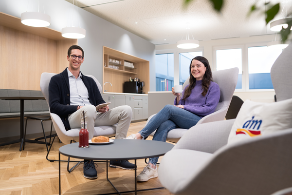 Zwei Mitarbeitende sitzen in einem modernen Café im Büro und unterhalten sich. Ein Mitarbeiter hält ein Tablet, während seine Kollegin eine Tasse Kaffee in der Hand hat. Das Café wirkt modern und hell.