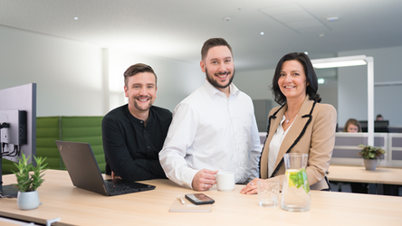 Zwei Mitarbeiter und eine Mitarbeiterin stehen nebeneinander vor einem höhenverstellbaren Schreibtisch im Büro und schauen in die Kamera. Der Mitarbeiter in der Mitte hält eine Kaffeetasse, während vor dem linken Mitarbeiter ein aufgeklappter Laptop steht.