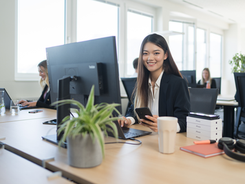 Eine lächelnde Mitarbeiterin sitzt an einem Schreibtisch in einem Büro und hält ein Telefon in der Hand. Vor ihr stehen ein Laptop mit separatem Bildschirm, eine Topfpflanze und Büromaterialien. In der Nähe arbeiten andere Mitarbeitende.