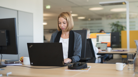 Eine Mitarbeiterin mit kurzen Haaren arbeitet konzentriert an einem Laptop in einem modernen Büro. Sie ist von Schreibtischen und Computern umgeben.
