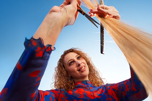 Eine Frau mit lockigem Haar, steht vor blauem Himmel schneidet langes blondes Haar. Dabei wirkt die Dame konzentriert und zufrieden.